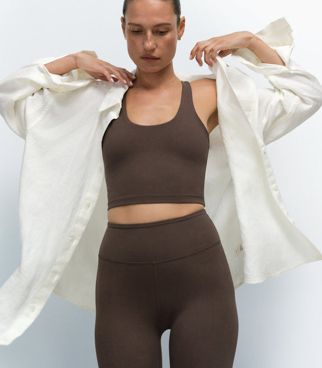 Model wearing a brown sports bra and matching leggings with a white denim jacket over the shoulders